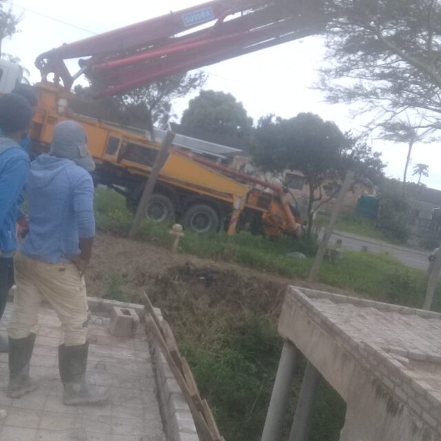 Yellow concrete boom pump truck on site with crew supervising, slab formwork visible in foreground