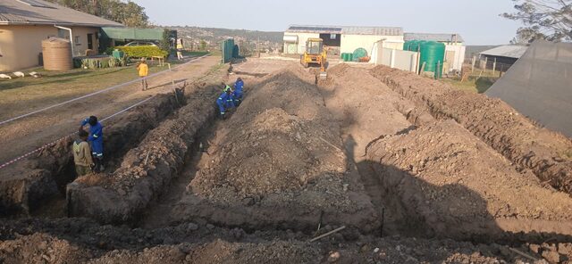 Wide twin trench excavation with crew in blue PPE overalls and yellow backhoe machine in the background on an Eastern Cape construction site