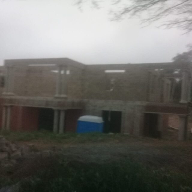 Double-storey structure mid-build showing brickwork and columns at upper floor level, rainy day on site