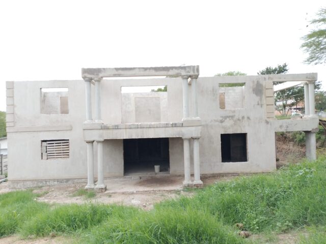 Near-complete double-storey plastered home with concrete columns and covered verandah in lush green surroundings
