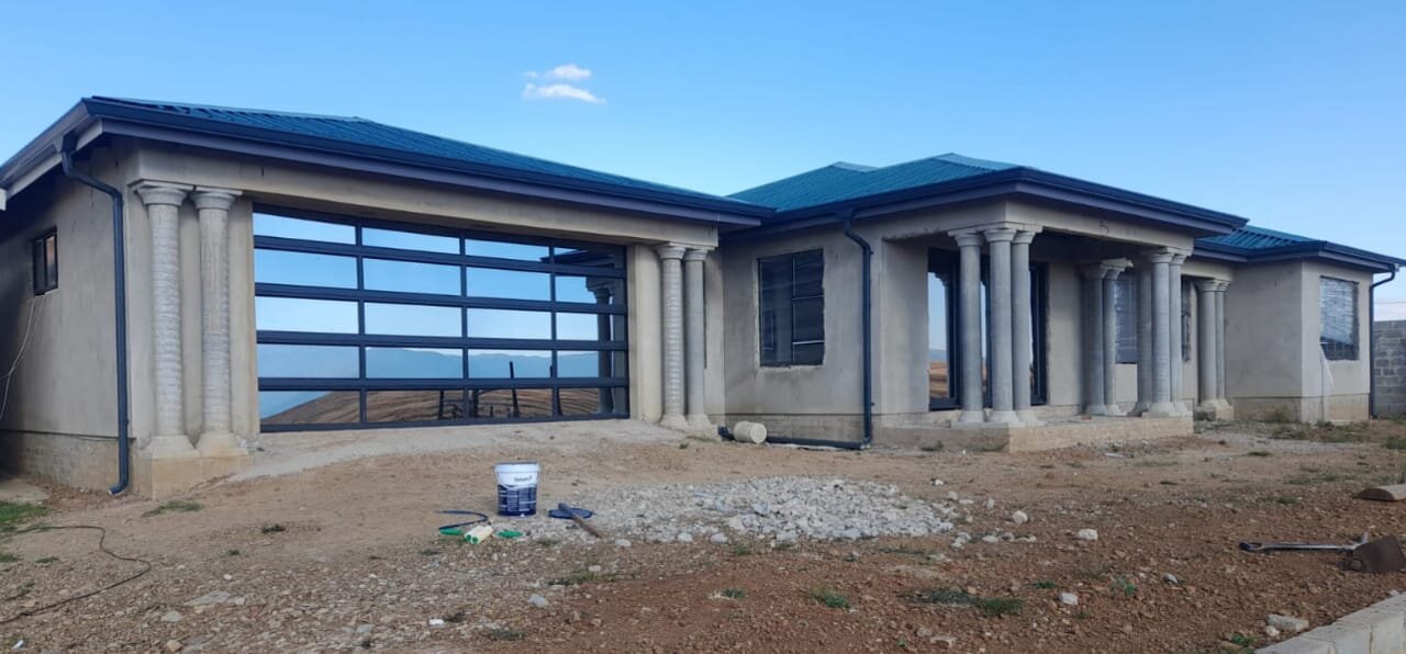 Completed single-storey home with concrete columns, dark teal metal roof, and glass roll-up garage door in Mount Ayliff, Eastern Cape